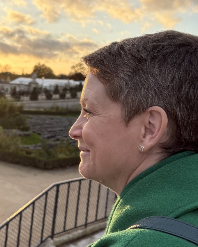 Jennifer Ellis in profile, smiling, looking out over a Paris garden at sunset in a green coat